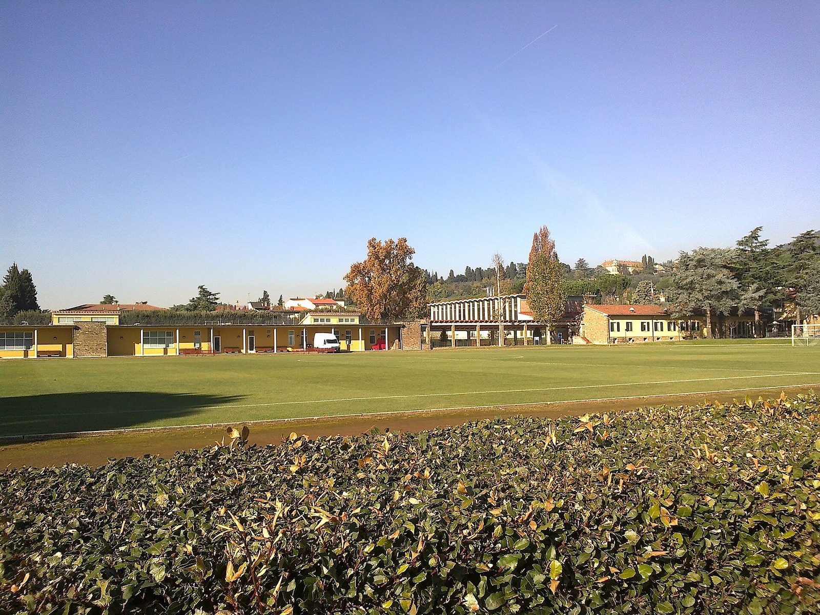 Un campo da calcio del Centro Tecnico Federale di Coverciano con gli edifici gialli del centro, alberi autunnali e le colline sullo sfondo