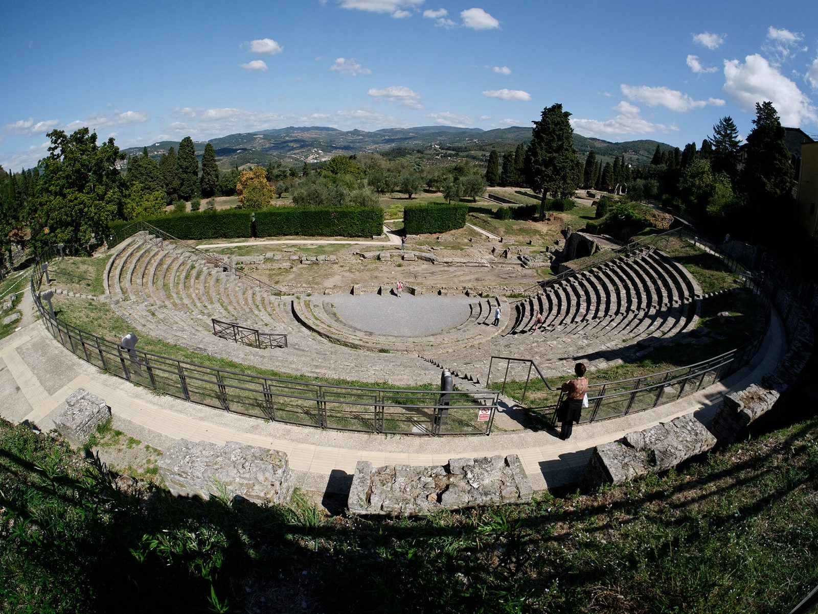 Il teatro romano di Fiesole visto dall'alto in una giornata di sole: la cavea semicircolare a gradoni in pietra quasi vuota, cipressi e ulivi intorno e le colline sullo sfondo sotto un cielo azzurro