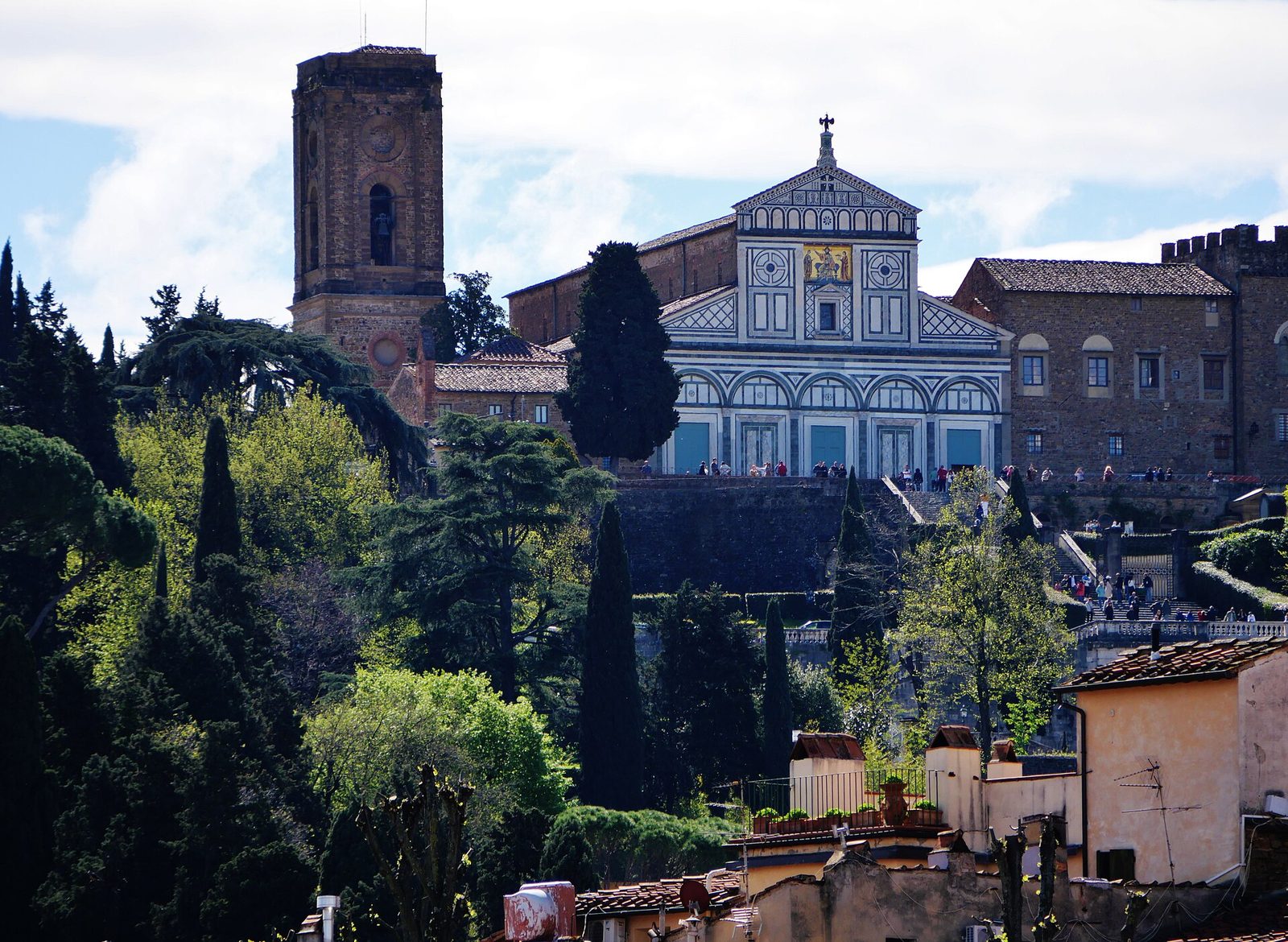 La facciata in marmo bianco e verde della basilica di San Miniato al Monte con il campanile in pietra, sulla collina tra cipressi e alberi, con tetti di case in primo piano