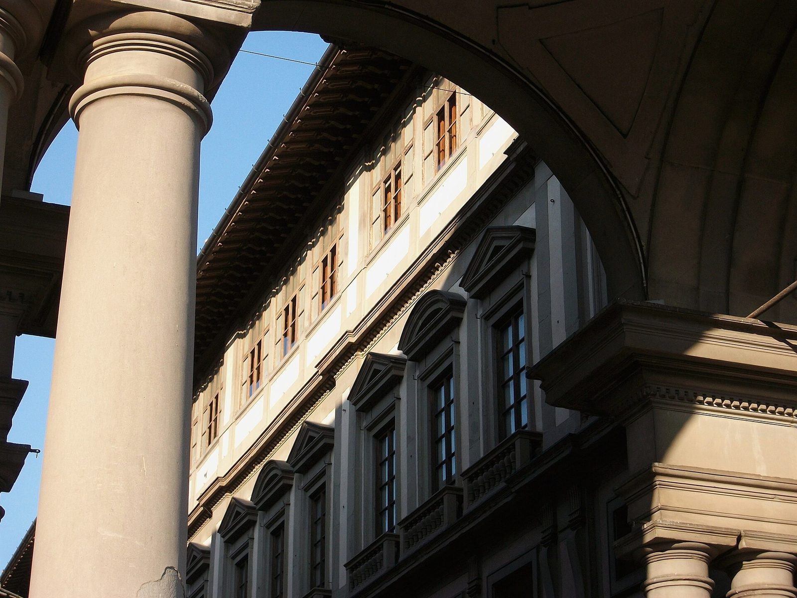 Una colonna e un arco del portico degli Uffizi incorniciano la facciata della Galleria, con le finestre dell'ultimo piano illuminate dal sole e il cielo azzurro